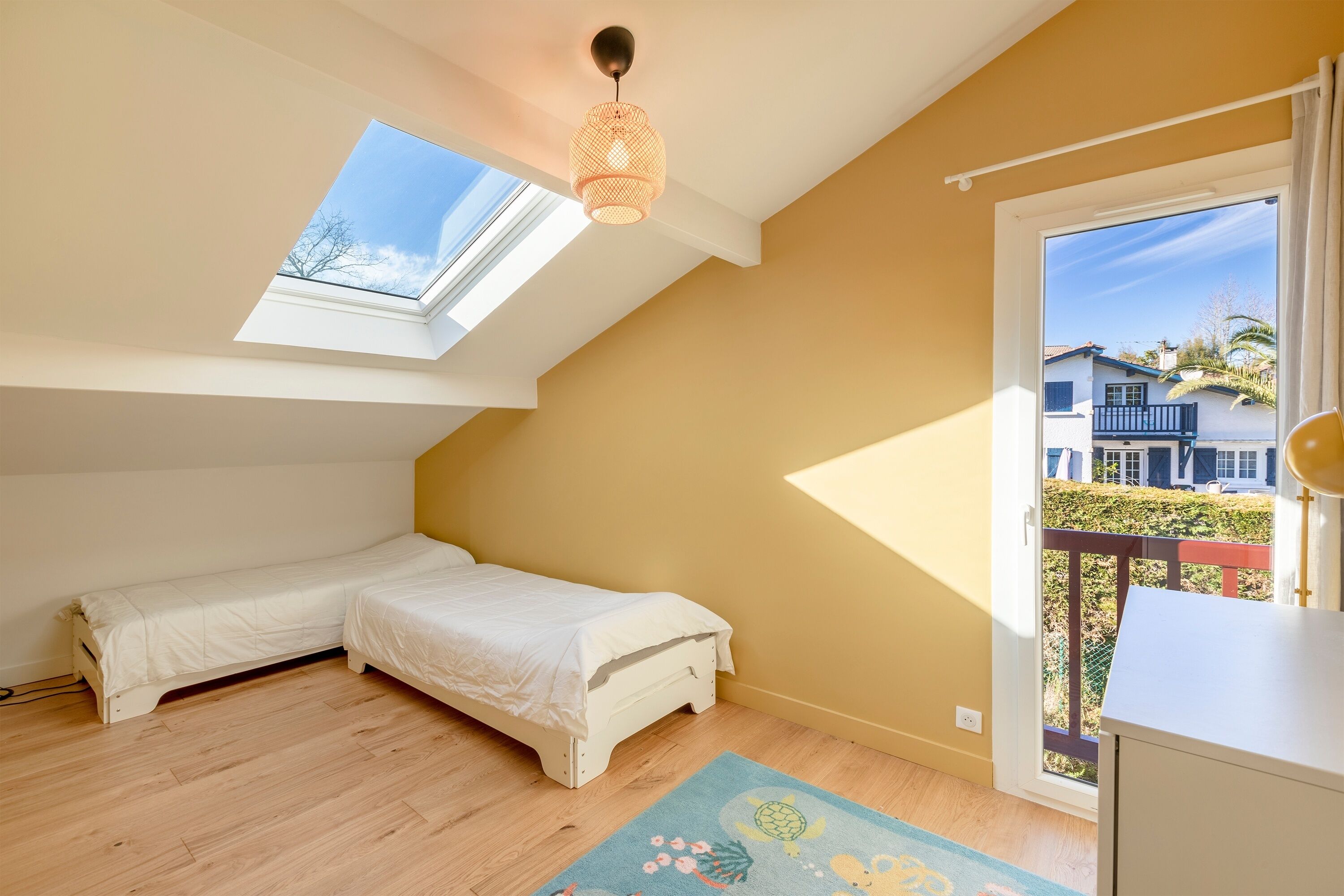 A bedroom with two single beds, a skylight, a balcony with a railing, and a view of houses.