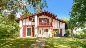 A two-story house with a red and white exterior, a stone pathway leading to the entrance, and a small patio with outdoor furniture. - La Senpere - Avec sauna entre Océan & Montagnes (Saint-Pée-sur-Nivelle)