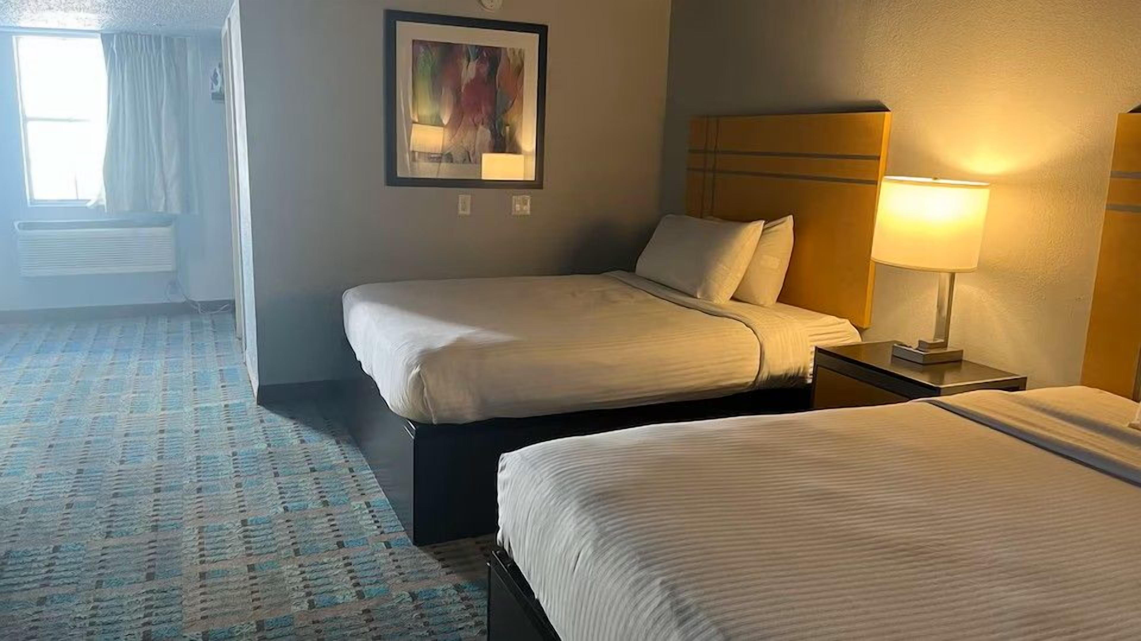 Quarto de hotel com duas camas, mesa de cabeceira com abajur, quadro emoldurado na parede e janela com cortinas.