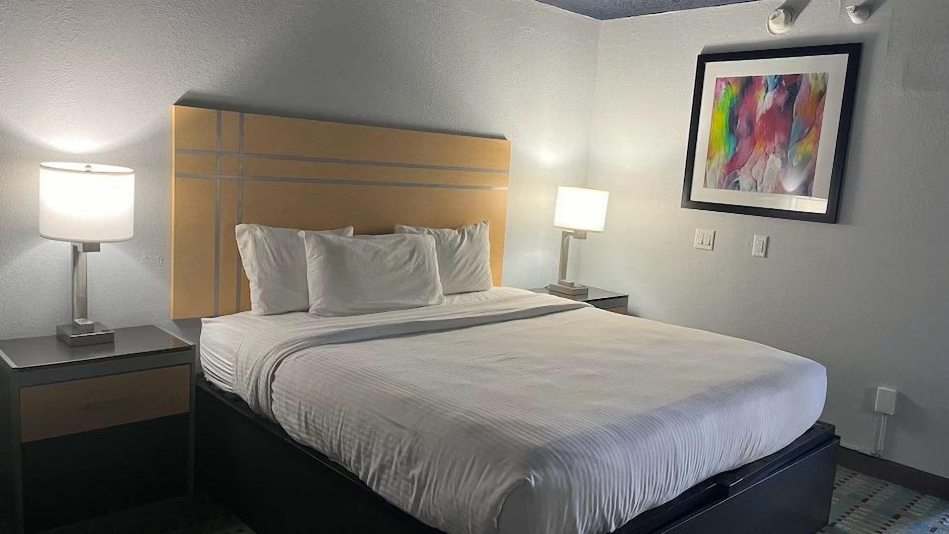 A hotel room with a bed, two bedside tables, lamps, a headboard, and a framed abstract painting on the wall.