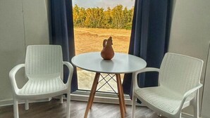 Et værelse med et rundt bord og to hvide stole, et vindue med blå gardiner, og udsigt til træer og himmel.