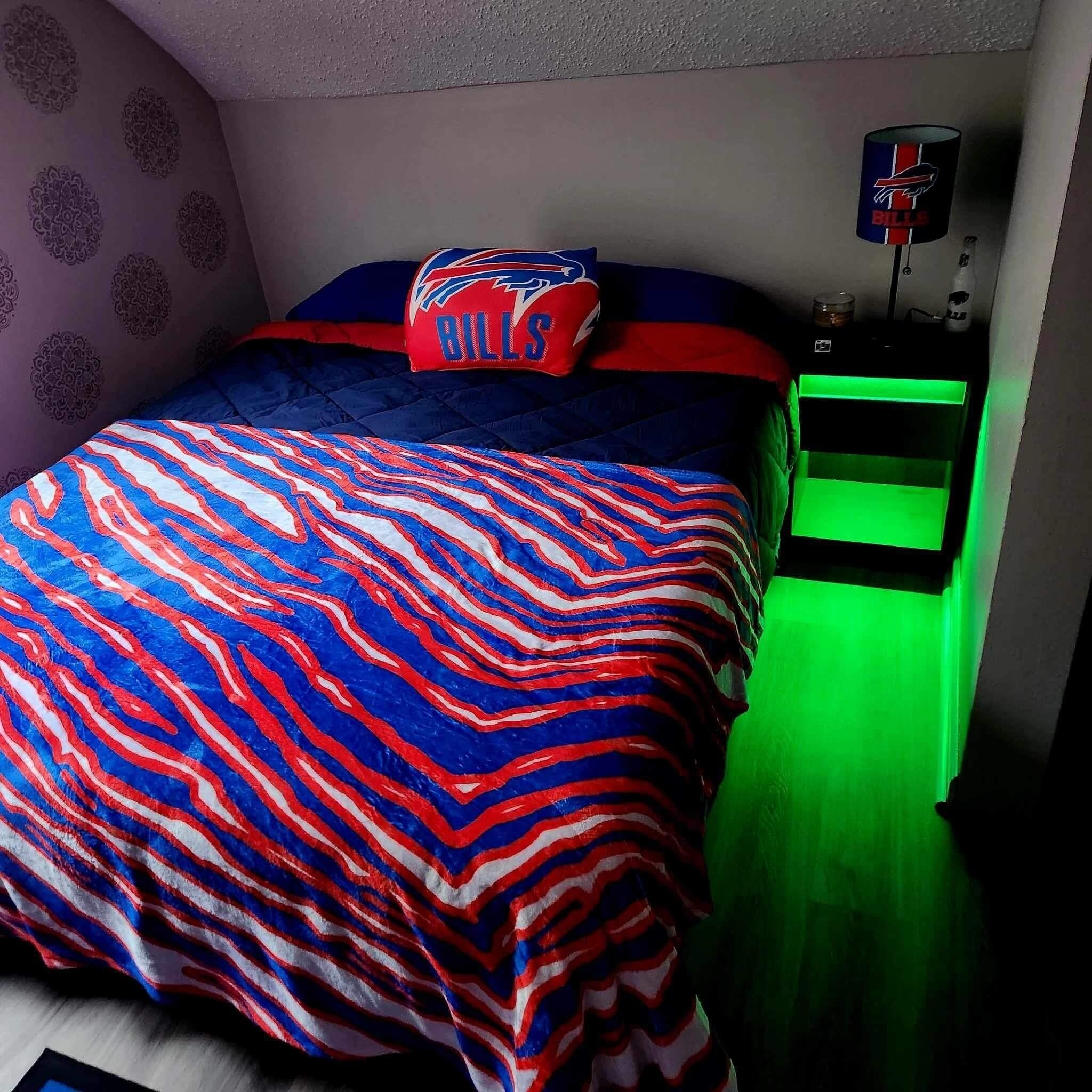 A bedroom with a bed, a pillow with a buffalo logo, a green-lit nightstand, and a wall with a circular pattern.
