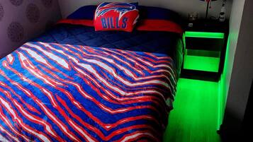A bedroom with a bed, a pillow with a buffalo logo, a green-lit nightstand, and a wall with a circular pattern.