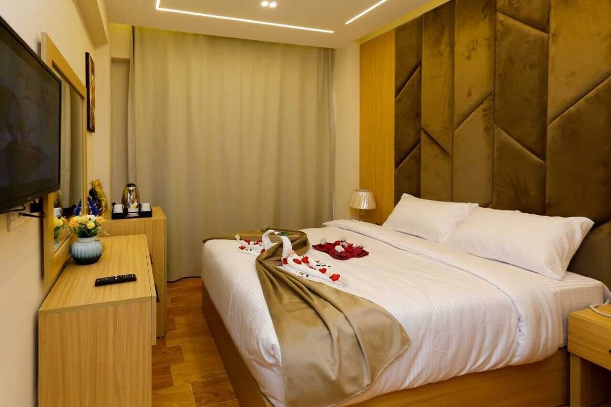 Habitación de hotel con cama, televisor, mesita de noche con un jarrón de flores y televisor montado en la pared.