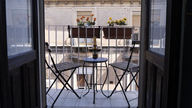 A balcony with two metal chairs and a small round table, a bottle of wine, and two flower pots with flowers.