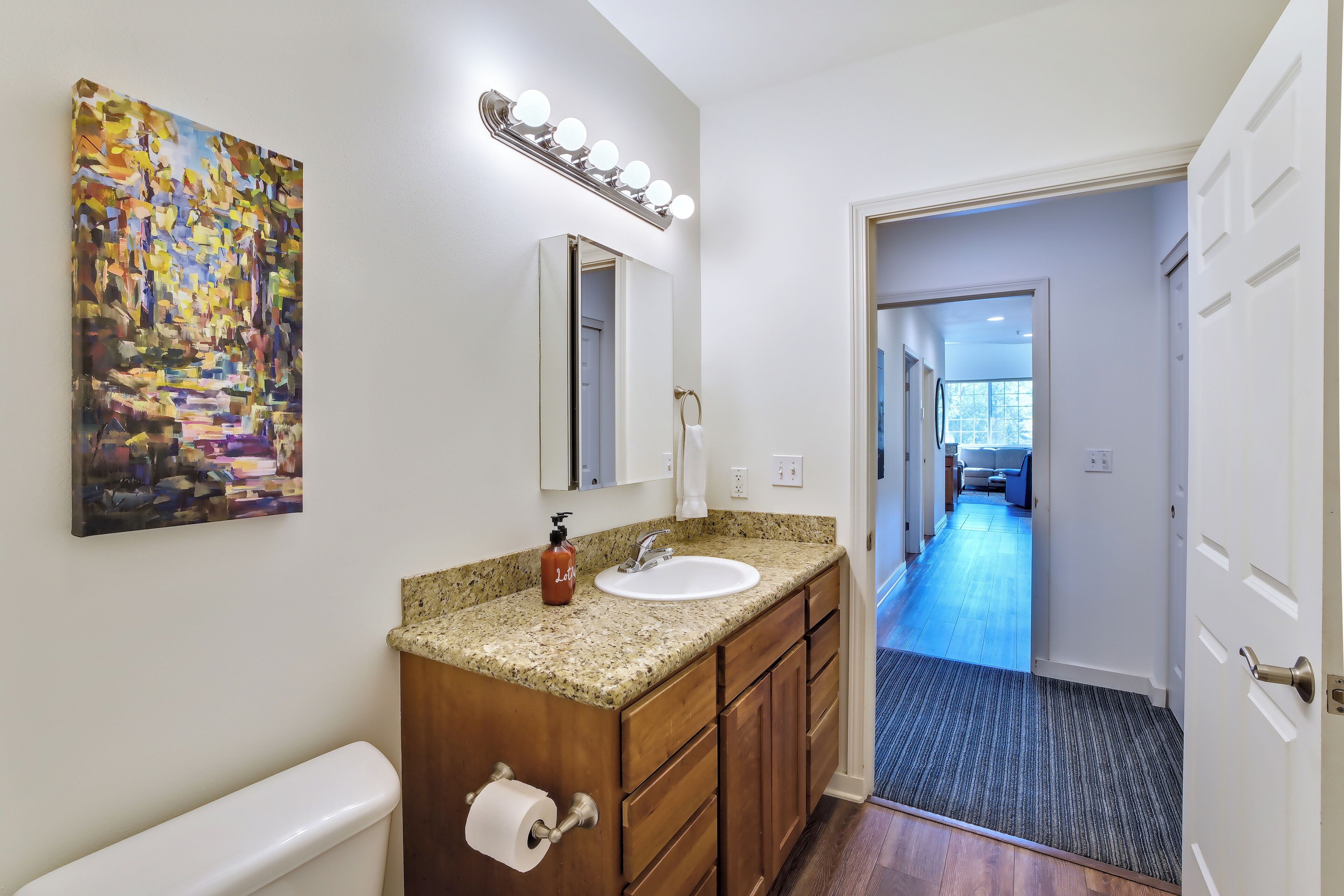 A bathroom with a granite countertop, a sink, a mirror with lights, a painting, and a hallway leading to a room.