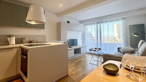 Une cuisine moderne dotée d’un îlot central, un coin repas avec une table dressée pour deux personnes, et un espace de vie comprenant un canapé et un téléviseur.