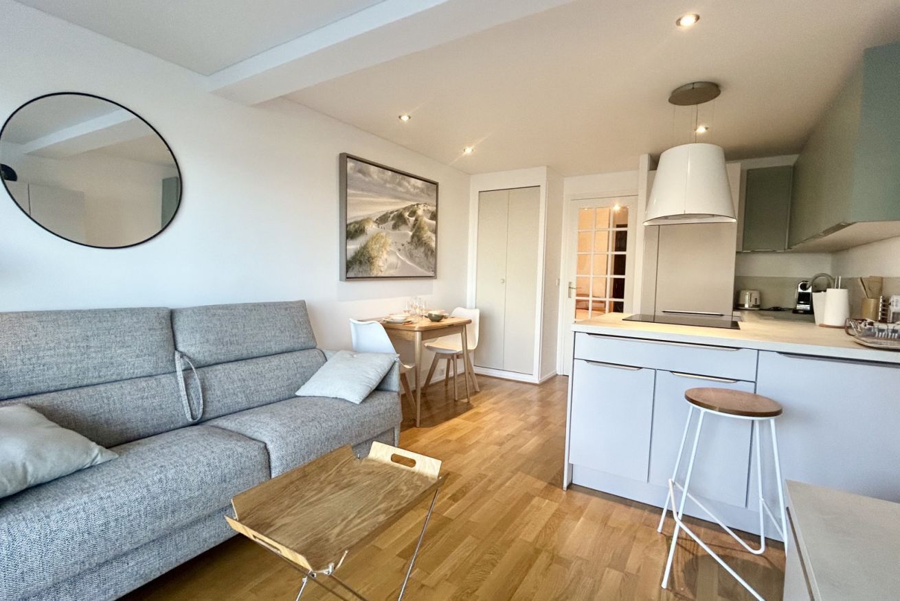 A modern living room with a grey sofa, wooden coffee table, and a dining area with a white table and chairs. A kitchen with a white island and bar stools is visible.