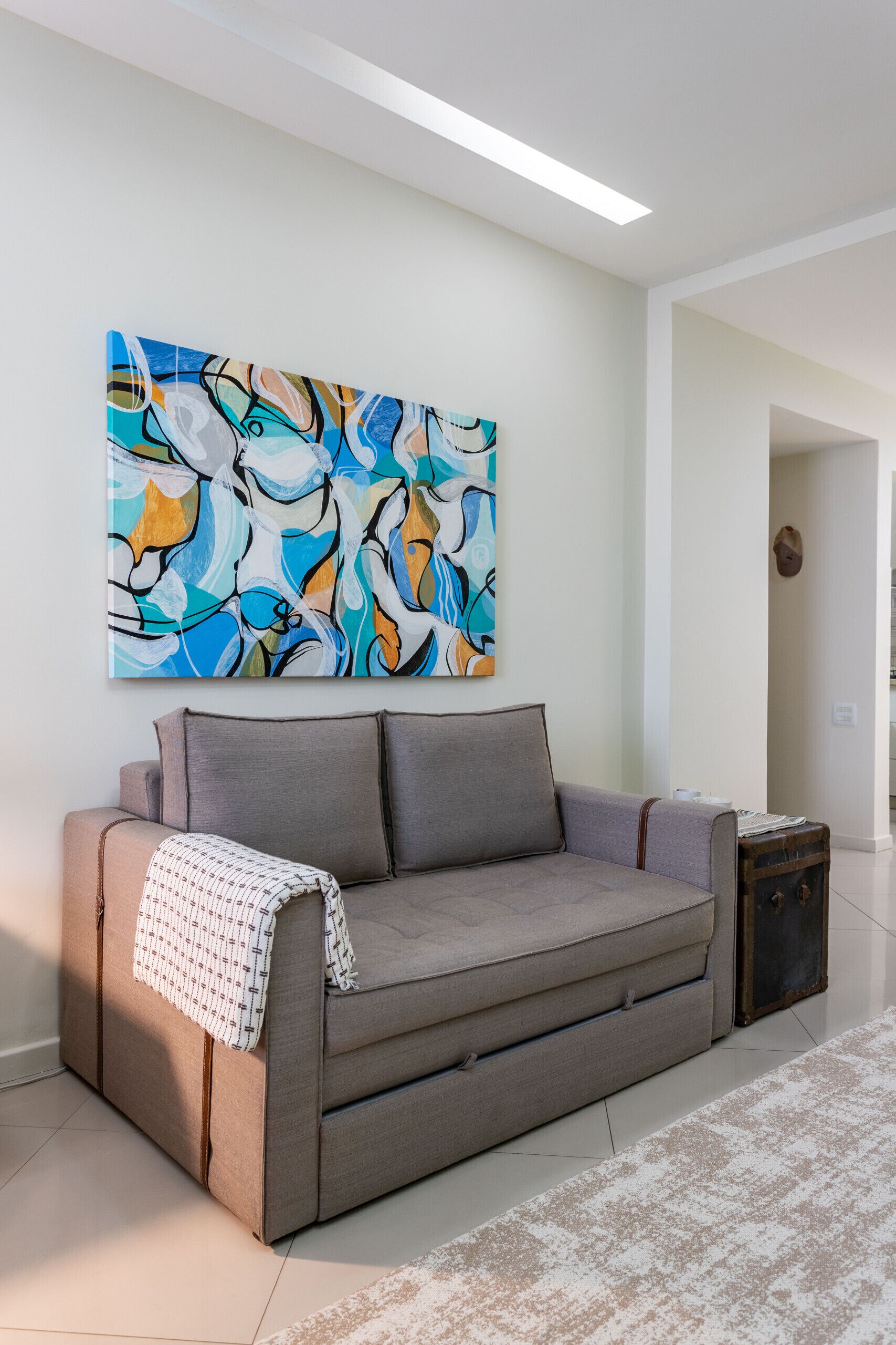 Uma sala de estar moderna com um sofá cinzento, uma pintura abstrata colorida e uma pequena mesa lateral de madeira.