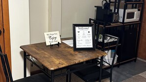 A small dining area with a wooden table and black chairs, a framed welcome sign, a clock, and a coffee station with a kettle, toaster, and microwave.