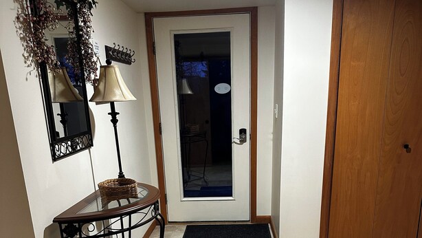 A hallway with a wooden door, a black doormat, a wicker basket on a console table, and a lamp.
