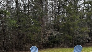 A fire pit surrounded by four Adirondack chairs in a grassy area with trees in the background.