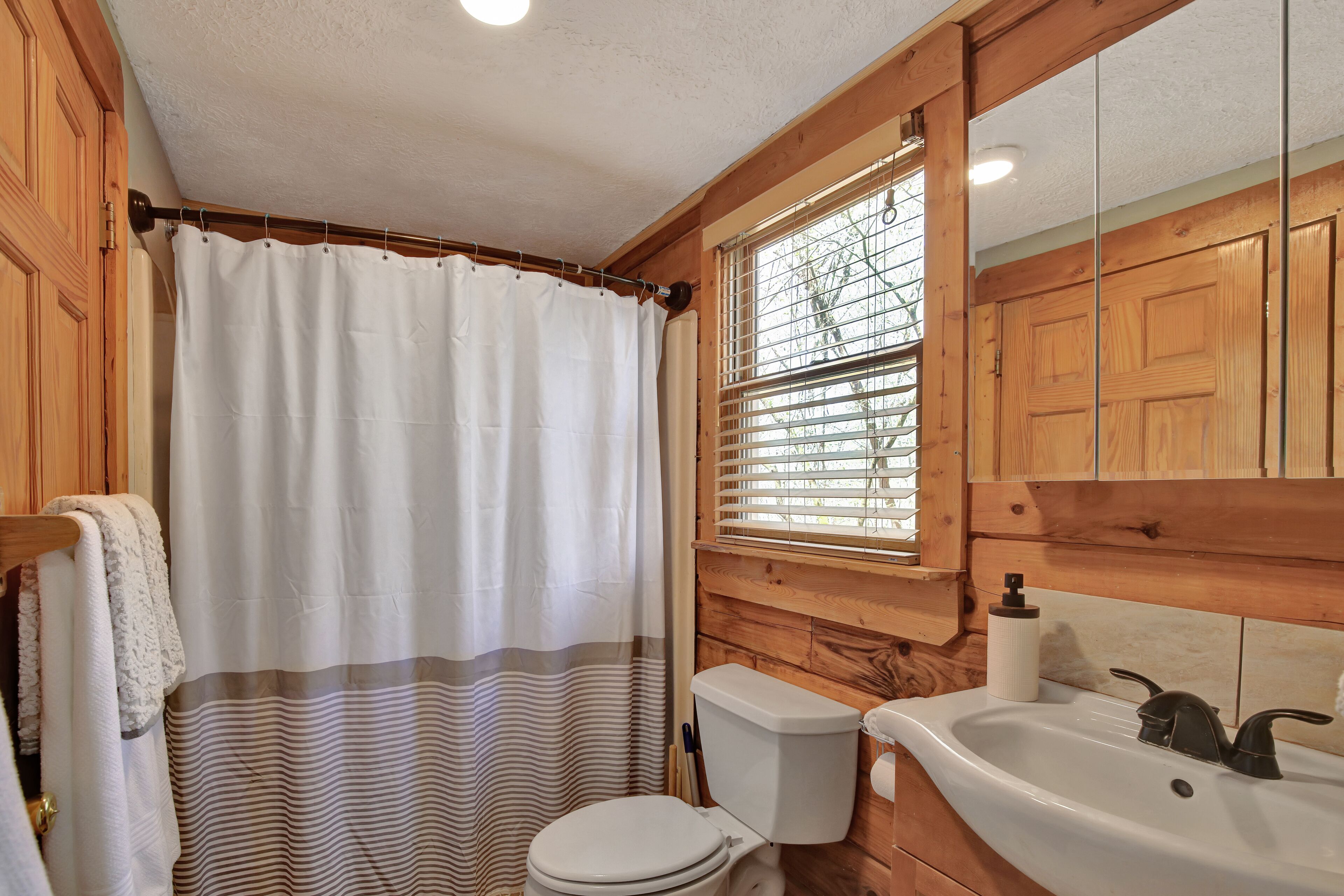 Un bagno in legno con tenda della doccia, WC, lavandino e finestra con persiane.