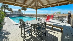 A covered pool area with a wooden deck, poolside chairs, and a view of the beach and buildings in the background.