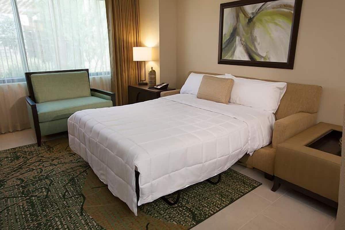 A hotel room with a bed, a chair, a nightstand, a lamp, and a painting on the wall.
