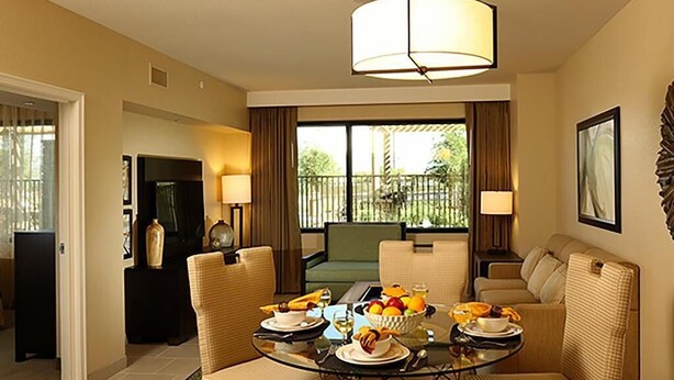 A dining area with a glass table, wicker chairs, and a bowl of fruit. There is a sofa and a television in the background. - The Berkley (Las Vegas)