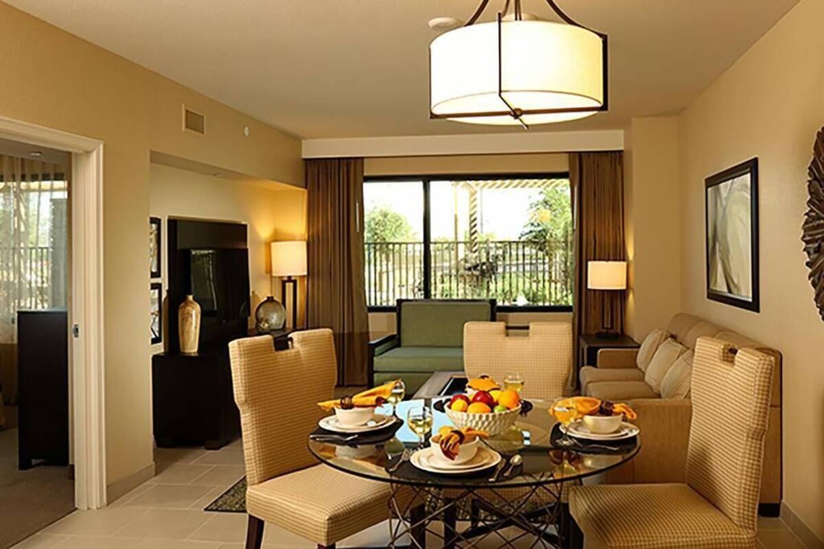 A dining area with a glass table, wicker chairs, and a bowl of fruit. There is a sofa and a television in the background.