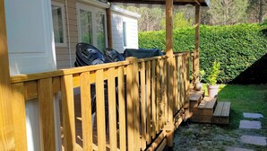 Un espacio al aire libre cubierto con una terraza de madera, un edificio blanco con ventanas y un seto verde al fondo.