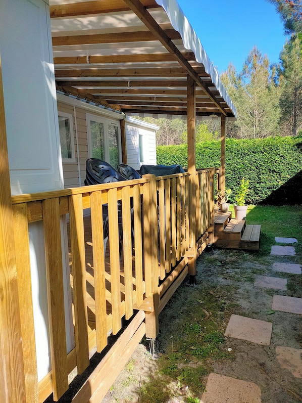 A covered outdoor area with a wooden deck, a white building with windows, and a green hedge in the background.