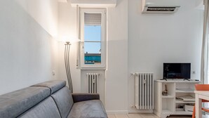 A modern living room with a grey sofa, a small table, a TV on a white cabinet, and a window with a view of a blue building.