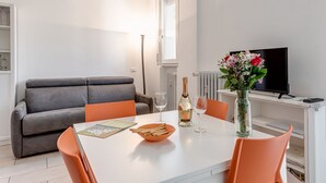 A modern living room with a grey sofa, a white dining table with orange chairs, a TV, and a vase with flowers.