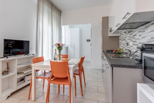 A modern kitchen with white cabinets, a countertop, and a built-in oven. There is a dining area with a white table and orange chairs.