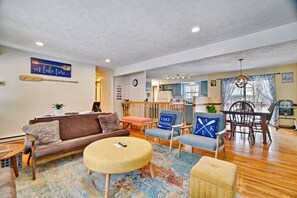 A cozy living room with a brown sofa, a round coffee table, and blue chairs with 'LAKE HOUSE' pillows. A kitchen is visible in the background. - Camp Cottontail Lakeside • Lakefront • Dock • Game Room • Firepit • Kayaks (Tobyhanna Township)