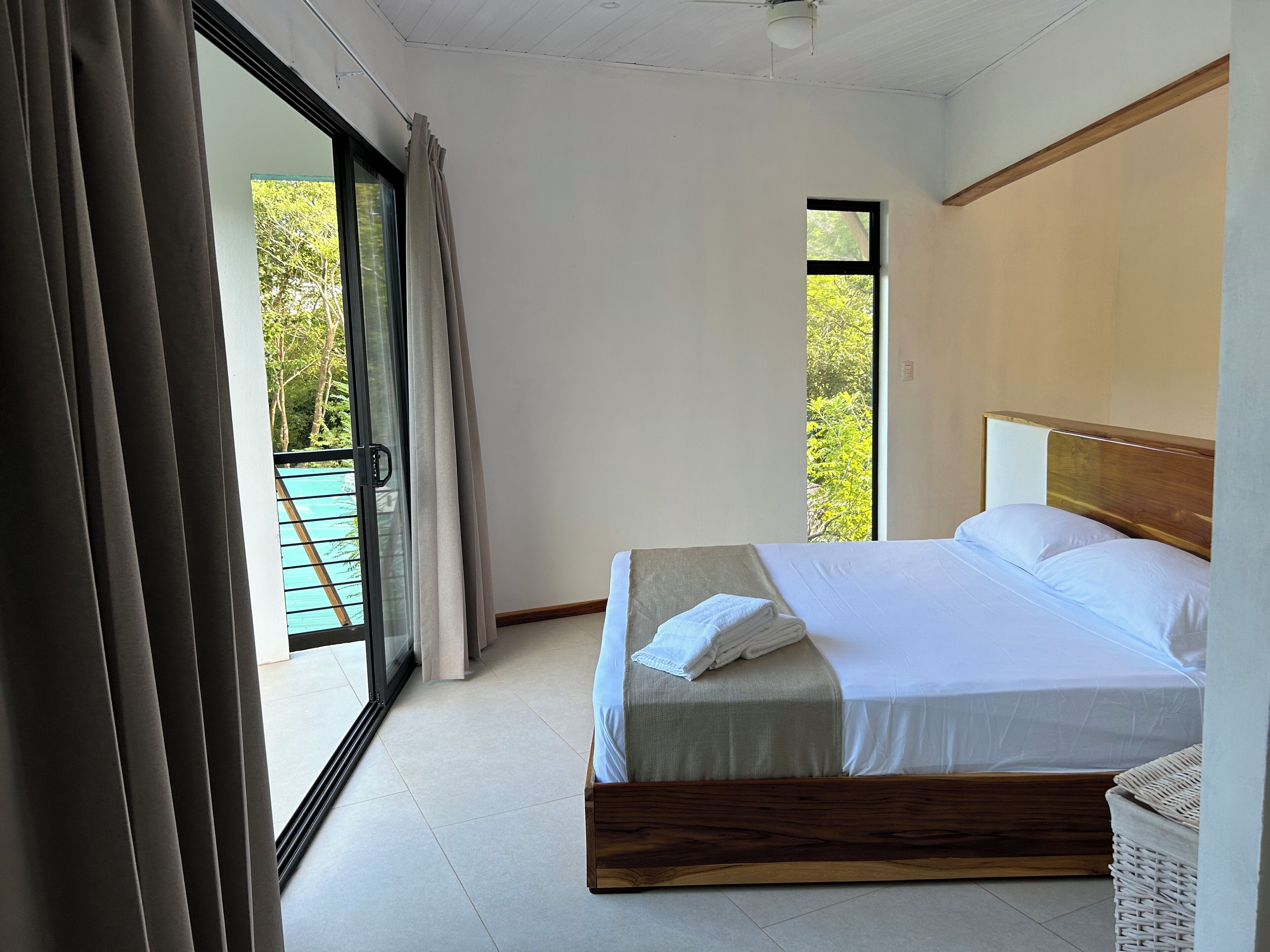 A hotel room with a large bed, a sliding glass door leading to a balcony, and a view of greenery outside.