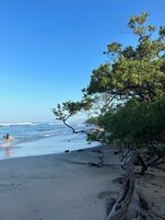 Una playa con cielos despejados, olas tranquilas y una persona caminando con una tabla de surf.