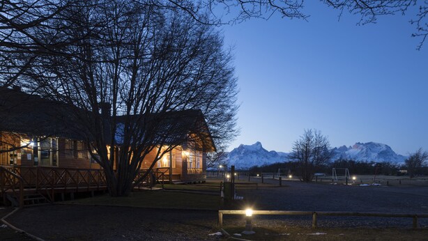 Une cabane en bois avec un toit de chaume, entourée d’un espace délimité par une clôture, avec une chaîne de montagnes en arrière-plan.