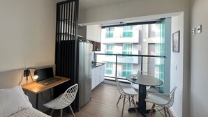 A modern, compact living space with a kitchenette, a dining area with a table and chairs, a bed with a white quilted cover, and a view of a residential building through large windows. - Studio em Perdizes - em frente a PUC (São Paulo)