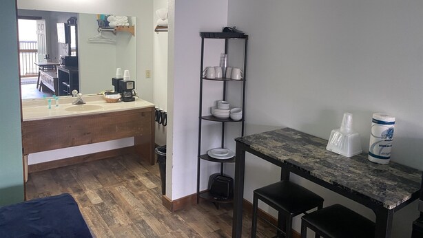 A compact room with a wooden vanity, a mirror, a shelf with cups and bowls, a small table with a roll of toilet paper, and a trash bin. - Double Queen Room w/Kitchen Supplies (Branson)