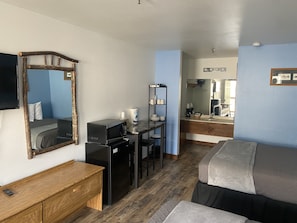 Une chambre d’hôtel compacte comprenant un lit, un bureau avec un ordinateur, un petit réfrigérateur, un four à micro-ondes, une étagère avec des tasses et une boîte de conserve, un miroir et une commode en bois.