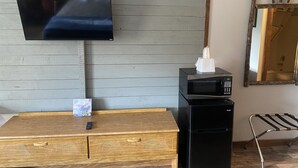 A room with a flat-screen TV mounted on a grey wall, a black microwave on a wooden table, a small bedside table with a remote control, and a mirror with a rustic frame. - Standard Double Queen Room (Branson)