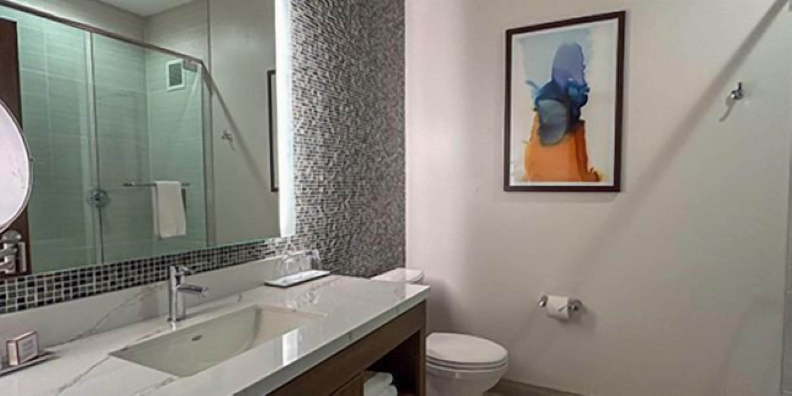 Une salle de bain avec un grand miroir, un lavabo, des toilettes et un tableau abstrait encadré accroché au mur.