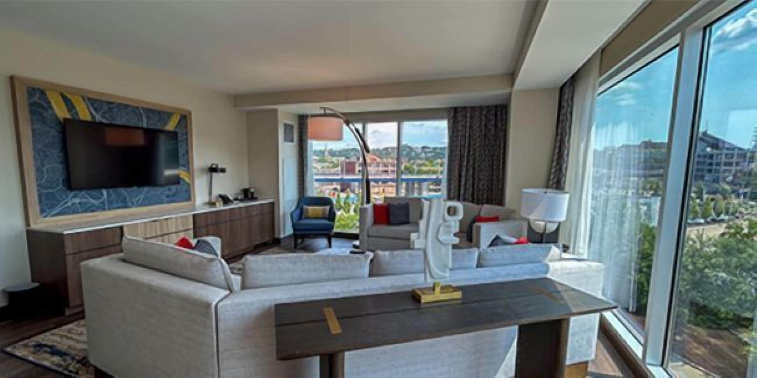 A modern living room with a large TV, a sofa, a coffee table, and a view of a cityscape through large windows.
