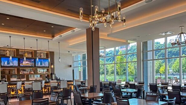 A spacious indoor area with multiple tables and chairs, a bar area with televisions, and large windows offering a view of greenery outside. - The Landing Hotel at Rivers Casino  (Pittsburgh)