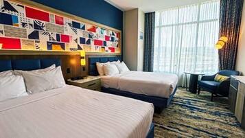 A hotel room with two beds, a large window with curtains, a wall art piece, and a small table with a chair.