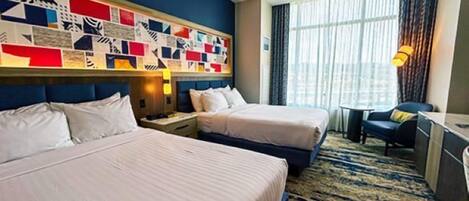 A hotel room with two beds, a large window with curtains, a wall art piece, and a small table with a chair.