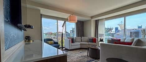 Habitación de hotel moderna con un ventanal grande que ofrece vistas a la ciudad, un sofá curvo, una pequeña mesa redonda y un escritorio con una lámpara.