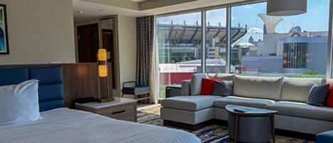 A modern hotel room with a large bed, a sofa, a coffee table, and a view of the cityscape through large windows.
