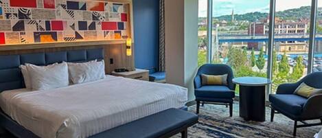 Une chambre d’hôtel avec un grand lit, deux fauteuils, une petite table et une vue sur la ville grâce à de grandes fenêtres.