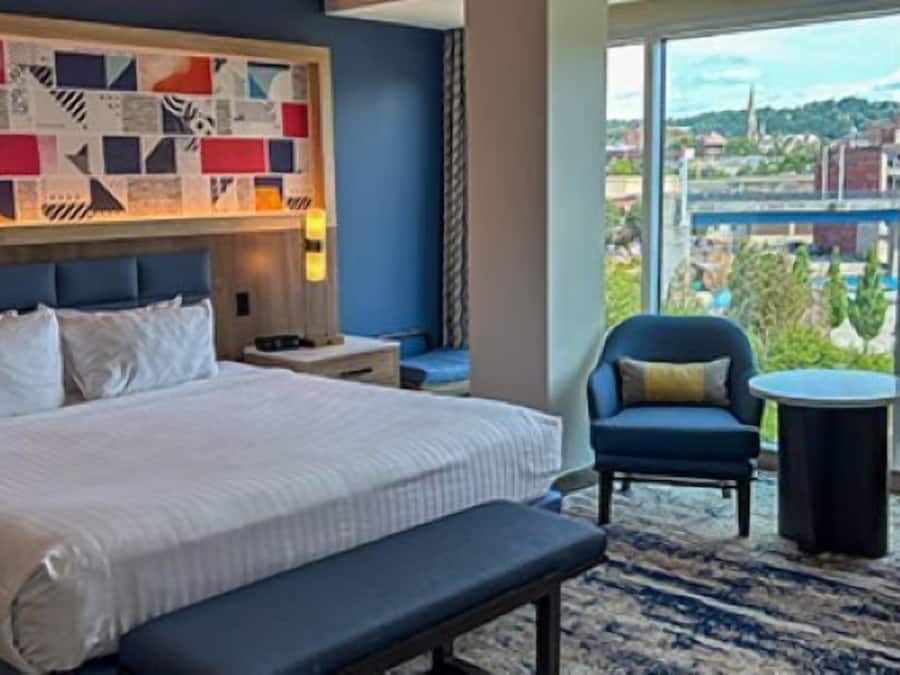 A hotel room with a large bed, two armchairs, a small table, and a view of the city through large windows.