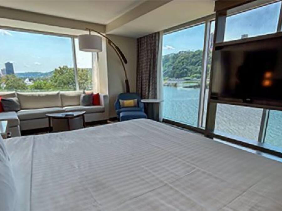 Quarto de hotel com uma cama grande, um sofá, uma mesinha e uma televisão. Há janelas amplas com vista para a água e edifícios.