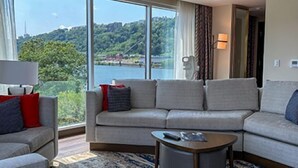 A modern living room with a large window offering a scenic view of a lake and hills. There is a sofa set and a small coffee table with a remote control on it. - The Landing Hotel at Rivers Casino  (Pittsburgh)