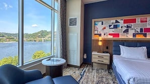 A hotel room with a large window offering a view of a body of water and greenery, a bed with white linens, a small round table, and a blue sofa. - The Landing Hotel at Rivers Casino  (Pittsburgh)