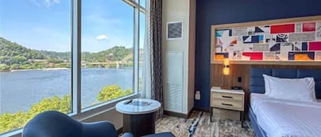 A hotel room with a large window offering a view of a body of water and greenery, a bed with white linens, a small round table, and a blue sofa.
