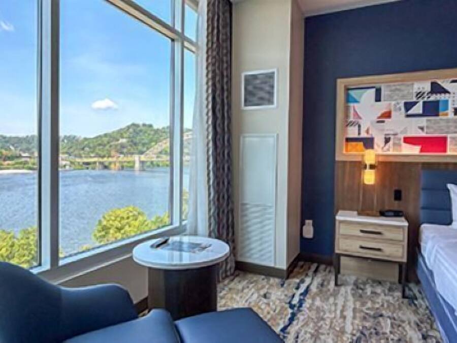 Habitación de hotel con un ventanal grande que ofrece vistas al agua y la vegetación, una cama con ropa blanca, una mesa redonda pequeña y un sofá azul.