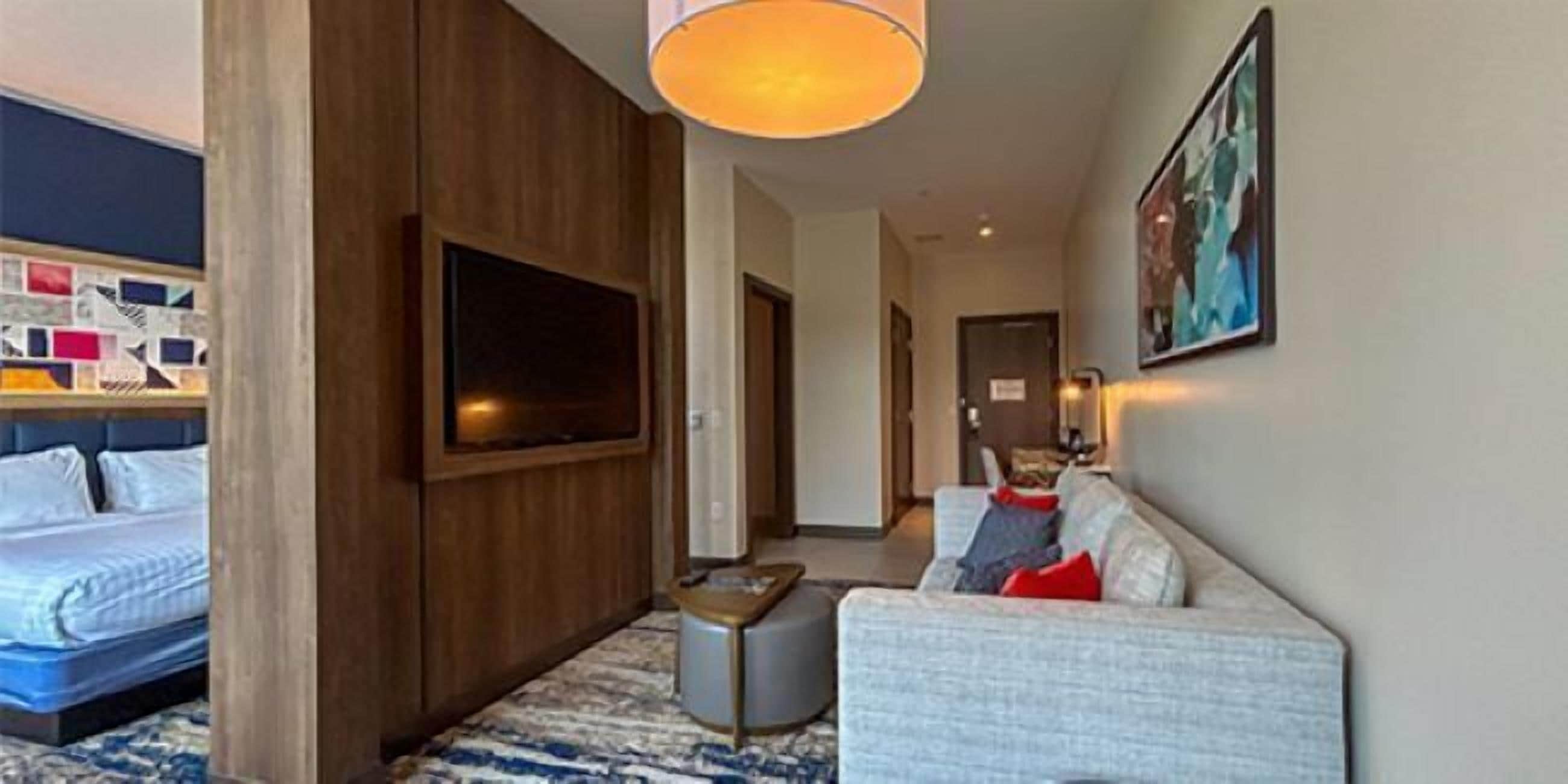 A hotel room with a bed, a TV mounted on the wall, a sofa, and a small ottoman.