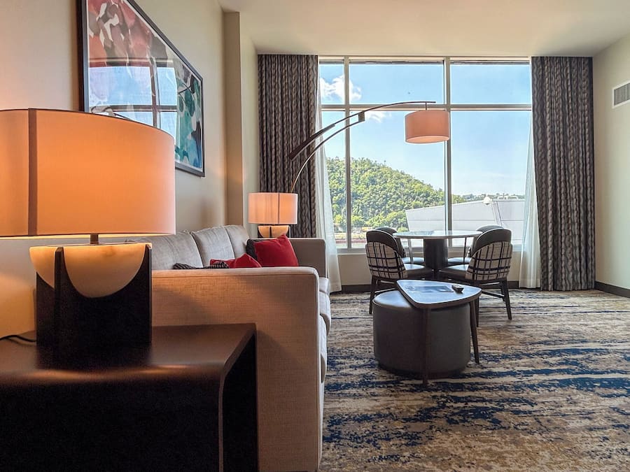 Une chambre d’hôtel avec un canapé, une petite table, une lampe, une chaise et une vue sur l’extérieur grâce à de grandes fenêtres.